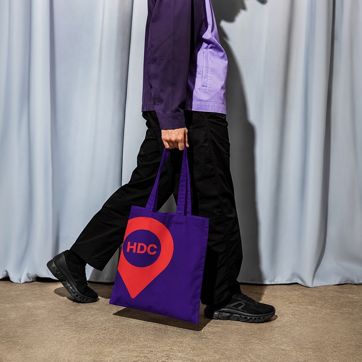 A person walking while carrying a purple Houston Design Collaborative tote bag.